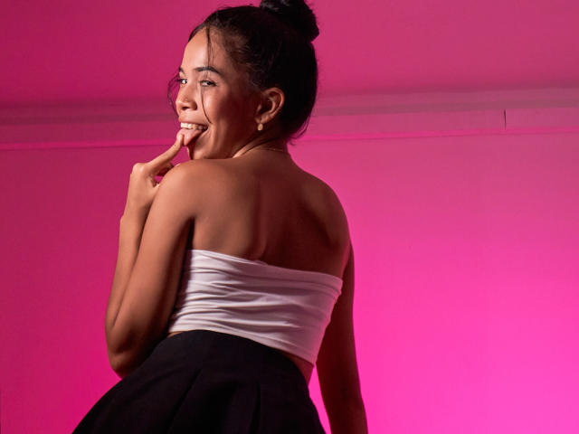 MarySmiith with her back to the camera, smiling, wearing a white strapless top and black skirt, her hand near her mouth, pink lighting, her hair in a bun.