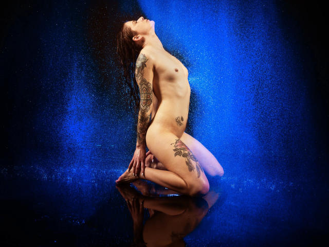 Nude Solveig-hot with tattoos, kneeling on wet floor, back arched, head tilted, water splashing, blue light, small tits, visible pussy, brown hair, tattoos on arm and thigh.