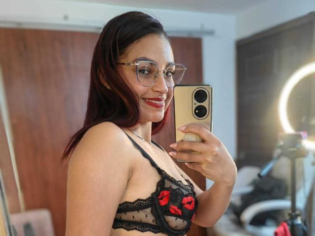 Ameliebx with red lipstick, glasses, and black lace bra with red kiss patterns, showing her tits in a mirror selfie, holding a gold phone, smiling.