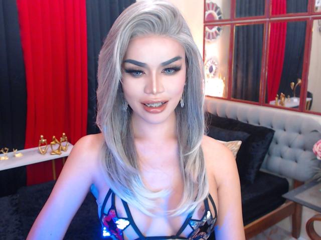 MindieMcleaks has silver hair, blue eyes, and is wearing a colorful strapless dress with visible tits. She's looking at the camera with a slight smile, showing a hint of her pussy.