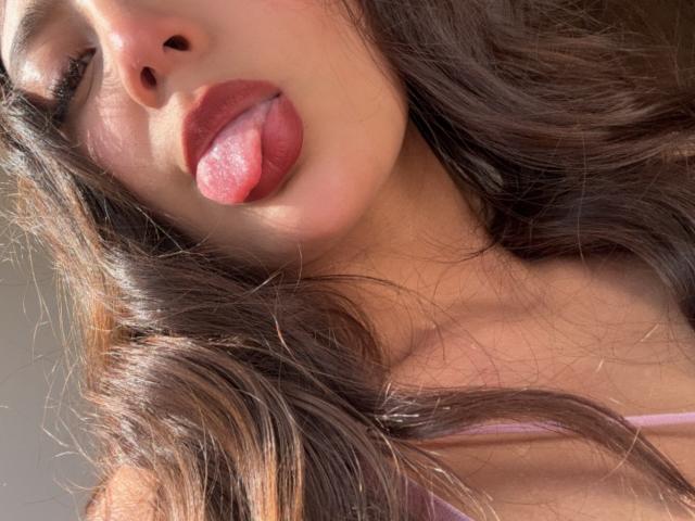 DianaFernandezz's face with her pink tongue sticking out, brown wavy hair, and slightly parted pink lips. Sunlight highlights her soft skin and pink top strap.