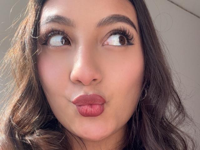 DianaFernandezz has pouty, pinkish-red lips, dark brown wavy hair, and brown eyes with long, thick eyelashes. She's looking to the side with a playful expression.