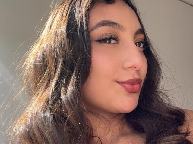 Das Gesicht von DianaFernandezz, mit welligem, braunem Haar, dunklem Eyeliner und rotem Lippenstift, zeigt ein leichtes Lächeln. Sie hat einen hellen Hautton und braune Augen. Ihre Brüste sind nicht sichtbar..