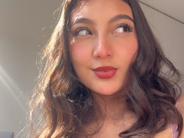 DianaFernandezz has wavy brown hair, full red lips, and dark, arched eyebrows. She's looking to the side with a subtle smile, sunlight highlighting her face.