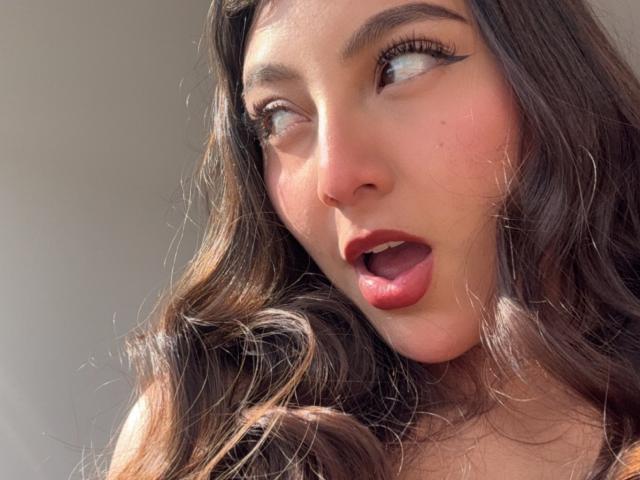 Het gezicht van DianaFernandezz, die naar boven kijkt met haar mond open, donkere eyeliner, golvend bruin haar, rode lippenstift en lichte huid, toont een verraste of opgewonden uitdrukking..