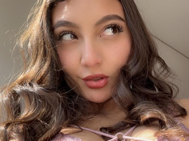 DianaFernandezz has wavy brown hair, full eyebrows, long lashes, and pink lips. Her brown eyes look to the side. She's wearing a pink lace top, showing her tits.