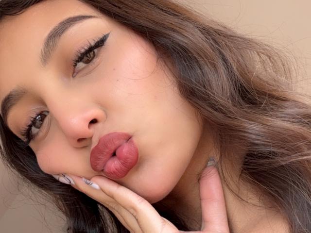 DianaFernandezz with pouty pink lips, brown wavy hair, and winged eyeliner, touching her cheek with fingers showing white and gray nail polish. Soft beige skin, brown eyes.