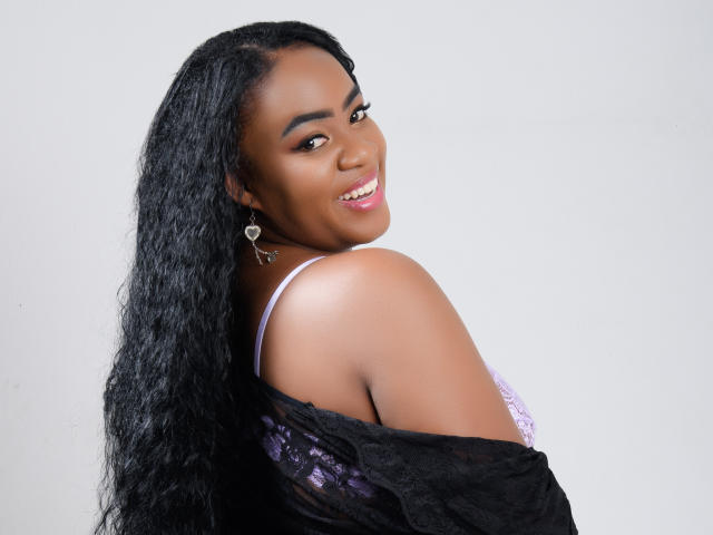 DanielaBrown with dark brown skin, long black wavy hair, smiling, looking over shoulder, wearing purple lace bra, black off-shoulder top, heart-shaped earrings, showing her tits.
