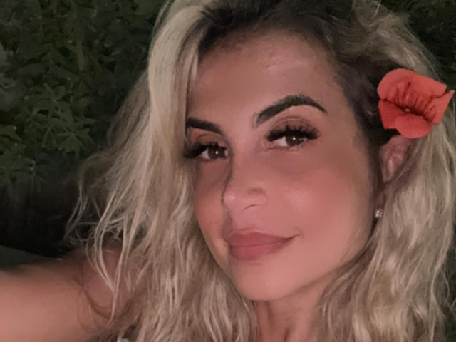Lellie has blonde wavy hair, a red flower in her hair, and a subtle smile with her tits slightly visible, showing off her cute face and full lips.
