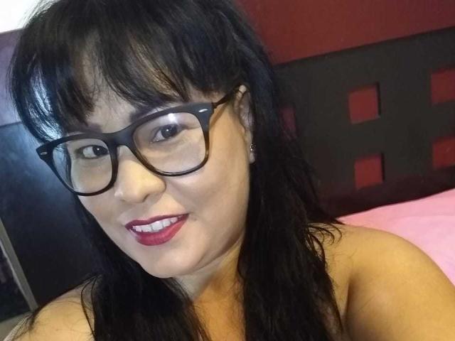 BetsyHottie with black glasses, red lipstick, and wavy black hair, smiling at the camera. She has cute earrings and is topless, showing her bare shoulders.