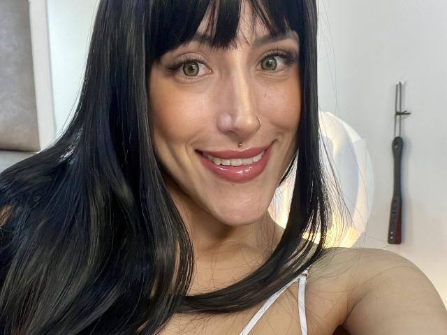 AlissaBrown has long black hair, green eyes, a nose ring, and a smiling face with glossy pink lips, wearing a white top, in a selfie.