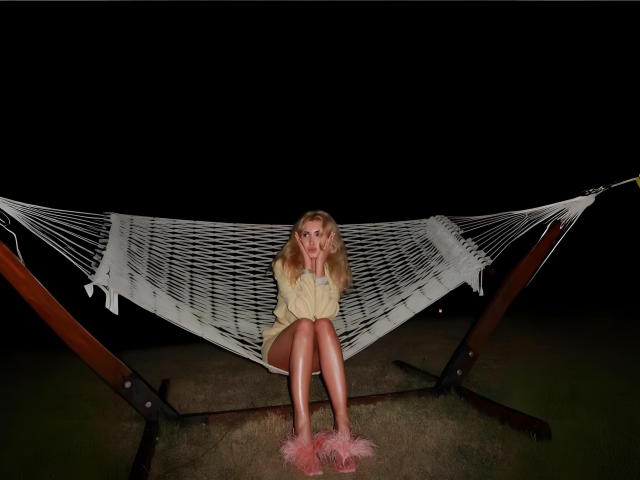 MashaTitova sits in a white hammock at night, wearing a yellow dress and pink fuzzy slippers, with her hand near her mouth, showing her tits and ass.