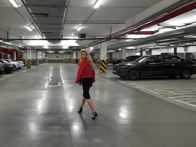 MashaTitova with long blonde hair, red top, black capri pants, and black high heels, walking in a parking garage, looking back over her shoulder, near a dark car.