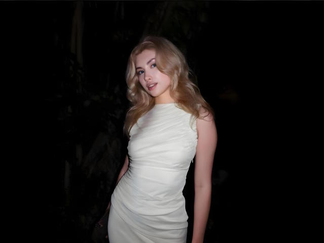 MashaTitova with blonde wavy hair, wearing a tight white dress, has small tits and a cute face, standing in dark with a slight smile, looking to the side.