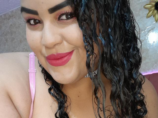 MelannieCarter with curly black hair, pink lipstick, and bold eyebrows, wearing a pink strap, smiling at the camera, showing her cleavage and a sparkling choker.