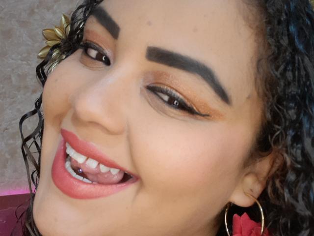 MelannieCarter's face, smiling with pink lips, showing teeth, brown eyes, curly black hair, bold eyebrows, orange eyeshadow, black eyeliner, gold flower hairpiece, and large red earrings.