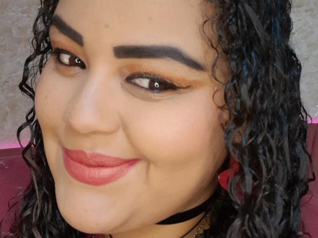 MelannieCarter has curly black hair, thick eyebrows, orange eyeshadow, winged eyeliner, red lipstick, and a cute smile. She's wearing a black choker. Her skin is light brown.