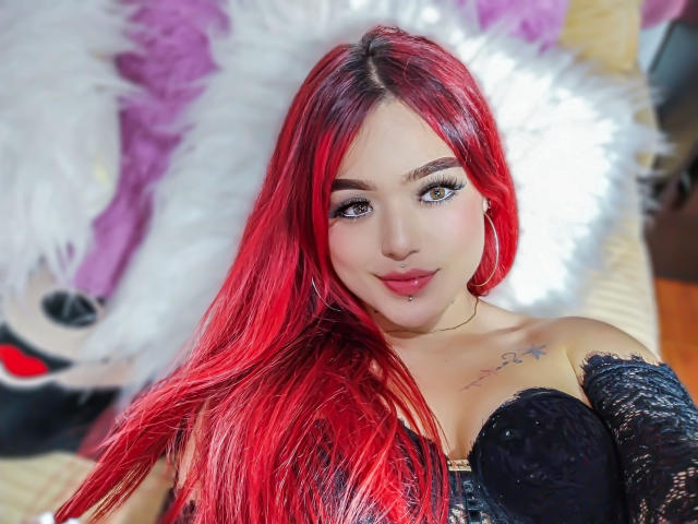 LeylakWith has bright red hair, blue eyes, and is wearing a black lace top. She has a nose piercing, lip piercing, and a small tattoo on her right chest.