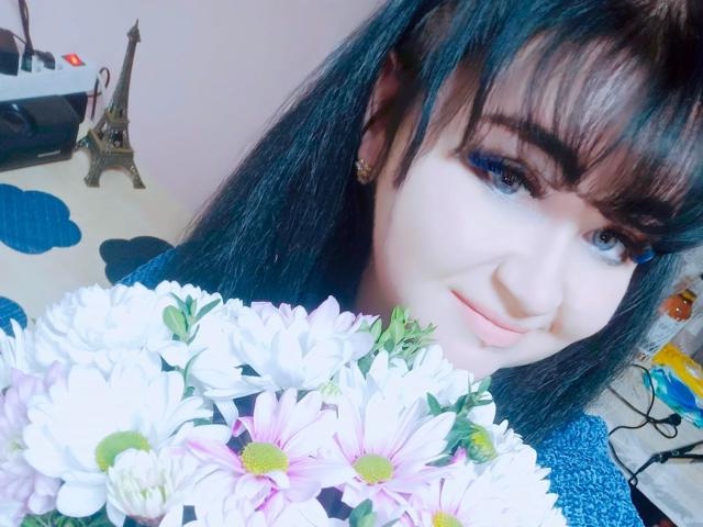 Kroox: Con i capelli blu-neri, la frangia e l'eyeliner blu, tiene un bouquet di margherite bianche e rosa, sorridendo leggermente. Foto ritagliata, pelle chiara, trucco leggero..