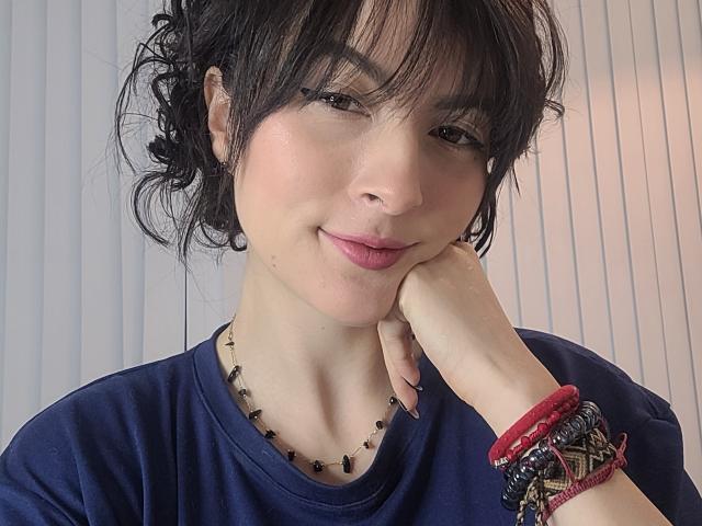 GabrielaMoore with black curly hair, blue shirt, bead necklace, red bracelets, hand on cheek, slight smile, fair skin, brown eyes, soft pink lips, looking at camera.