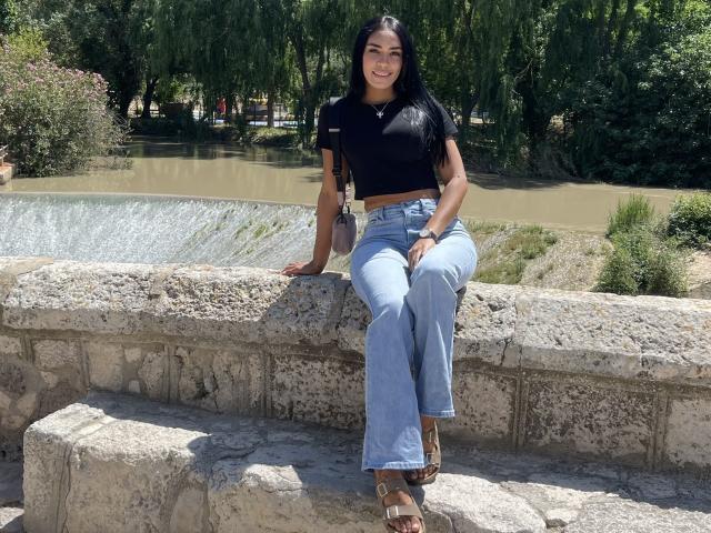 KylieeSex, with long black hair and a smile, sits on a stone wall in blue jeans and a black crop top, showing her taut midriff and brown sandals.