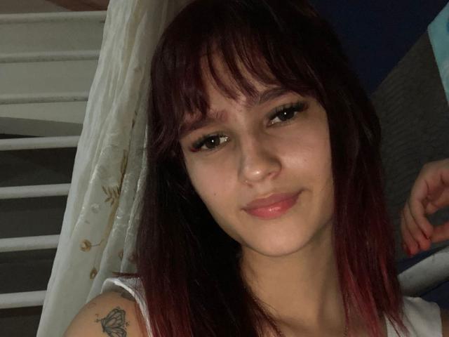MelodyMansson has dark red hair with bangs, a small tattoo on her right arm, and is smiling with pink lips. Her brown eyes are slightly open.
