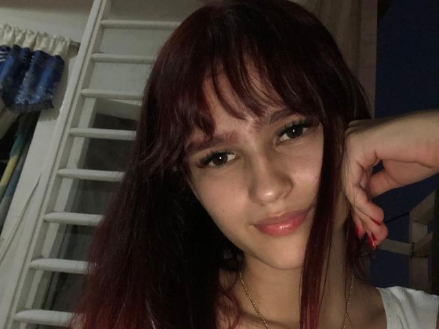MelodyMansson has dark red hair, brown eyes, and a small beauty mark on her right cheek. She's smiling with a hand on her head, wearing a white top.