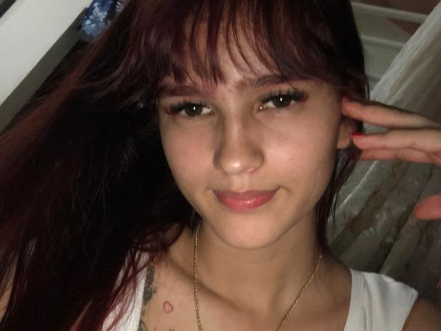 MelodyMansson has a cute face with dark brown hair, green eyes, and a small smile. She's wearing a white top, a gold necklace, and has a small tattoo on her chest.