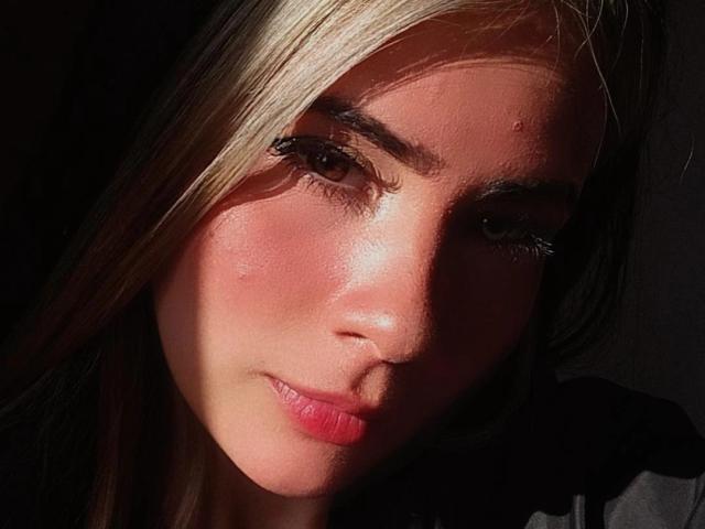 KrystinBrowm's face with sunlit blonde hair, pink lips, flushed cheeks, and a small pimple on her nose, dark shadowed eyes, and subtle smile.