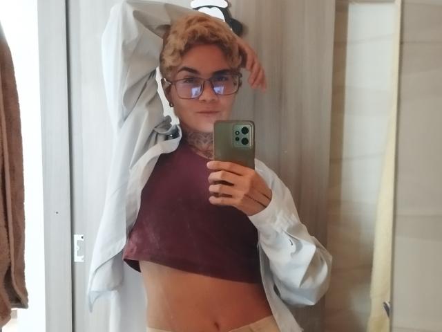 AnaRoa mit kurzen, lockigen, blonden Haaren, einer Brille und einem Tattoo unter ihrem Kinn, die ein Telefon hält, ein aufgeknöpftes Hemd und ein burgunderfarbenes Crop-Top trägt, wobei ihre kleinen Brüste sichtbar sind..