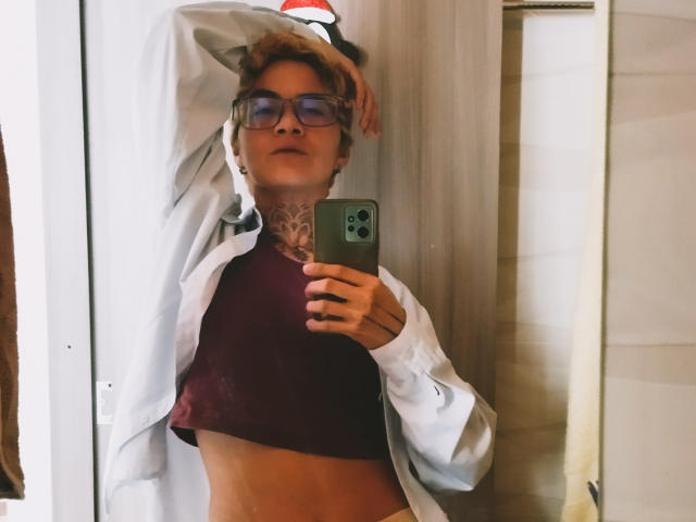 AnaRoa, con gafas, cabello castaño corto y tatuajes, vestida con una camisa blanca abierta y una blusa burdeos, tomándose un selfie en el espejo, mostrando su abdomen y su teléfono con una funda verde..