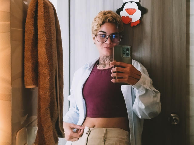 AnaRoa, avec ses cheveux blonds bouclés, ses lunettes, son tatouage au cou, un haut bordeaux, une chemise ouverte, un pantalon beige, tenant un téléphone, un selfie devant un miroir, une serviette marron, un autocollant de pingouin, souriante, se reflétant dans la porte..