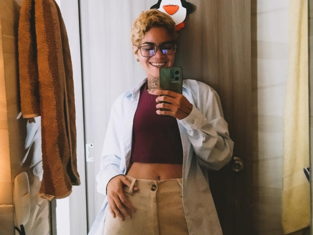 AnaRoa, met krullend blond haar, een bril en een glimlach, draagt een rode crop top, witte broek en een open blauw shirt, en maakt een spiegelzelfportret met een groene telefoon..