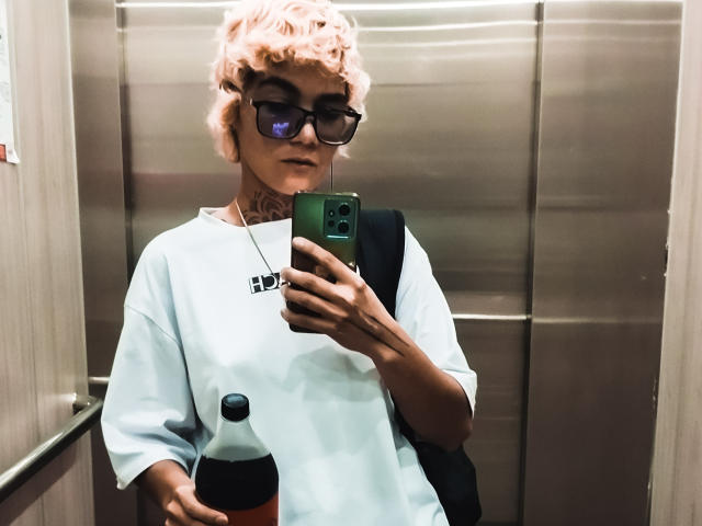AnaRoa, con i capelli ricci rosa, gli occhiali neri e una maglietta bianca, tiene il telefono in ascensore, con uno zaino nero, una collana e una bottiglia arancione in mano. Si scatta un selfie riflesso nello specchio..