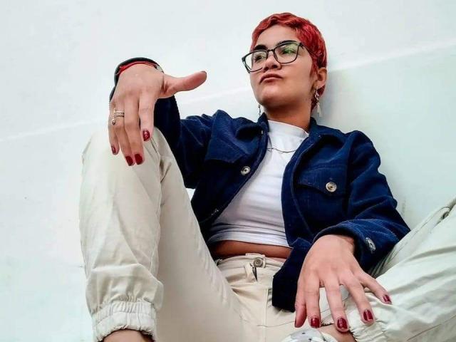 AnaRoa with red hair, glasses, and red nails, wearing a denim jacket, white tee, and beige pants, sitting with one leg up, hand pointing, ring on finger.