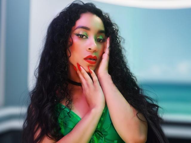 KylieJen met lang, krullend zwart haar, groene oogschaduw, rode lippenstift en rode nagels, die haar gezicht aanraakt, met een groene top, een zwarte choker, en de oceaan op de achtergrond..