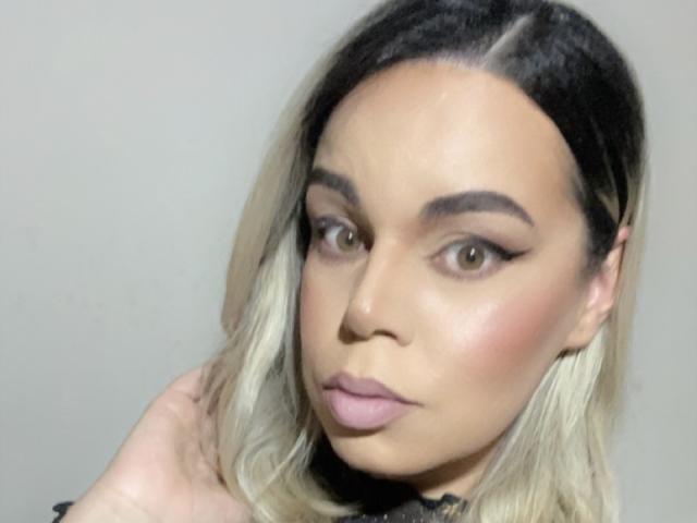 FernandaPassiva tem cabelos com um degradê de loiro para preto, olhos castanhos claros e lábios rosados, com uma expressão séria. Suas sobrancelhas são grossas e ela está tocando o cabelo com a mão..
