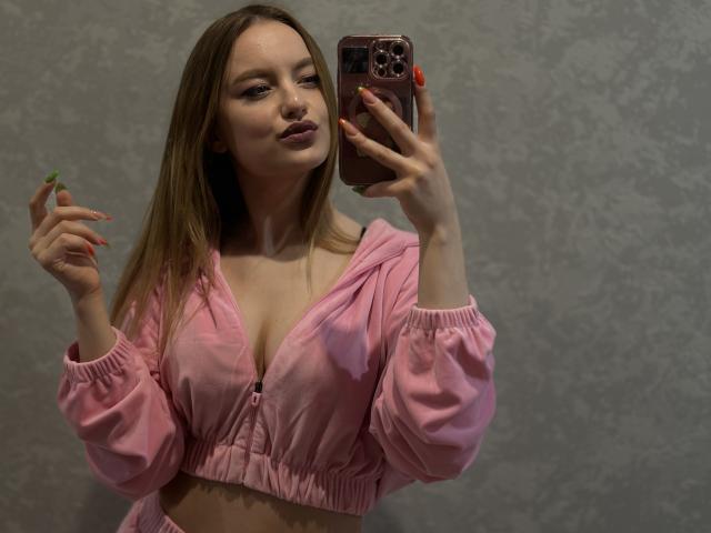 SweetOhGirl with long brown hair, pink puffy jacket, and cleavage, holding phone with pink case, pink nails, and green ring, looking at mirror, smirking.