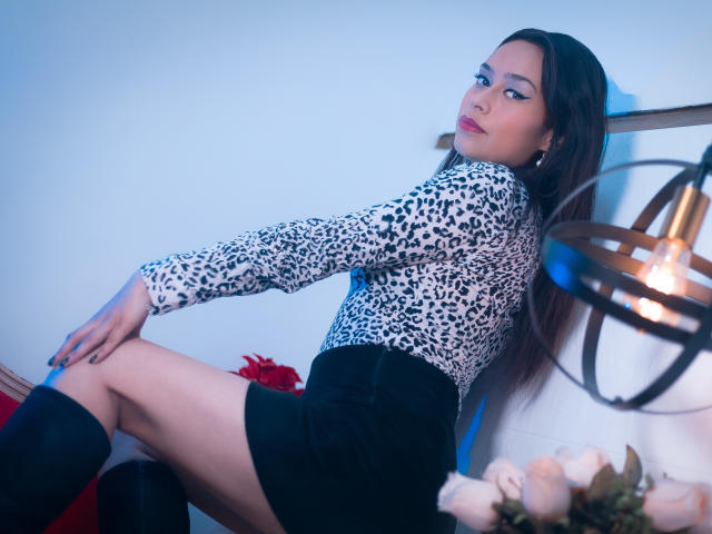 Felicity Fate mit langem, dunklem Haar, die ein Top mit Leopardenmuster und schwarze Shorts trägt, sitzt mit einem Bein angezogen, blickt über ihre Schulter und trägt schwarze Kniestrümpfe..