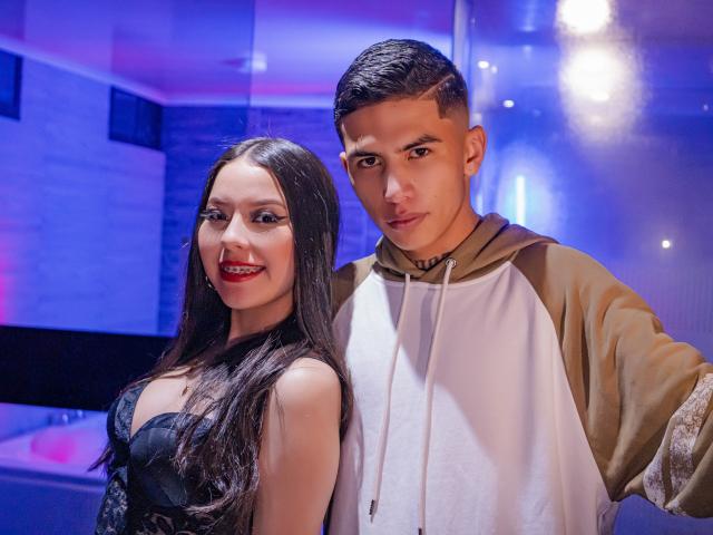 KylieAndJustin with long black hair and red lipstick, wearing a black lace top, stands next to Justin with short brown hair, wearing a beige and white hoodie.