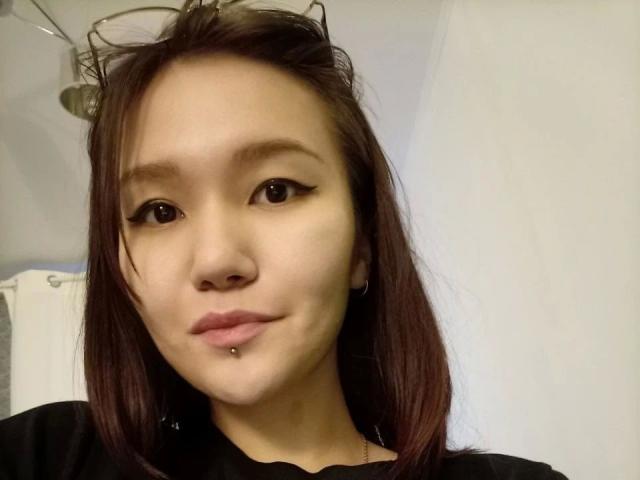 Kelly-Yon-hot has light brown skin, straight brown hair, black eyeliner, a nose piercing, and a lower lip piercing. She's wearing a black shirt and has glasses on her head.