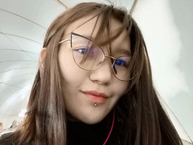 Kelly-Yon-hot with brown hair, glasses, blue eye, purple eye, nose piercing, and black turtleneck, looking cute. Her lip piercing is visible, and her hair is slightly messy.