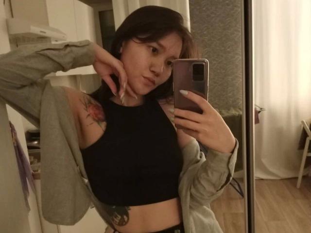 Kelly-Yon-hot with shoulder tattoo, black sports bra, gray jacket, holding phone, mirror selfie, short brown hair, pouty expression, visible ass tattoo, wooden floor, dim lighting.
