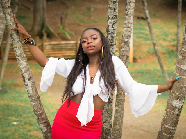SamyLorenss with dark skin, long black hair, white tied-up blouse, red high-waist skirt, pink eyeshadow, standing between trees, holding branches, wearing black bracelet, red lipstick.