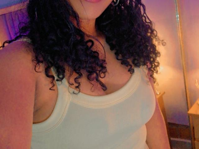 JackelineRoset heeft krullend zwart haar en draagt een witte, geribbelde tanktop die haar borsten laat zien. Ze staat op een plek met een warme, gele achtergrond. Haar huid heeft een medium tint..