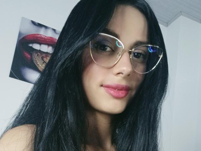 AnaisHilton with long black hair, wearing gold-rimmed glasses, pink lipstick, and a slight smile, showing off her cute face and sexy, subtle features.
