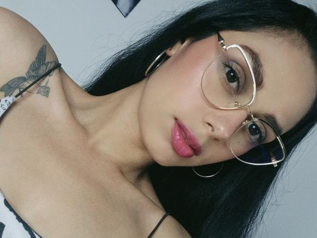 AnaisHilton with long black hair, wearing gold-rimmed glasses, hoop earrings, pink lipstick, and a black strappy top, showing a bird tattoo on her right shoulder.