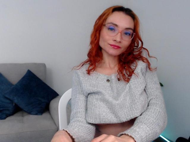 Drikka, con i capelli rossi ricci, gli occhiali e una maglia grigia a maglia, seduta su una sedia bianca, che mostra il seno, con cuscini blu su un divano grigio..