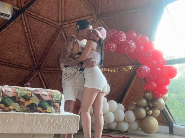 LucyAndJake, a tattooed dude in a white tank and shorts, kisses a hot chick in a pink cap and white shorts, her ass in his hands, balloons behind.