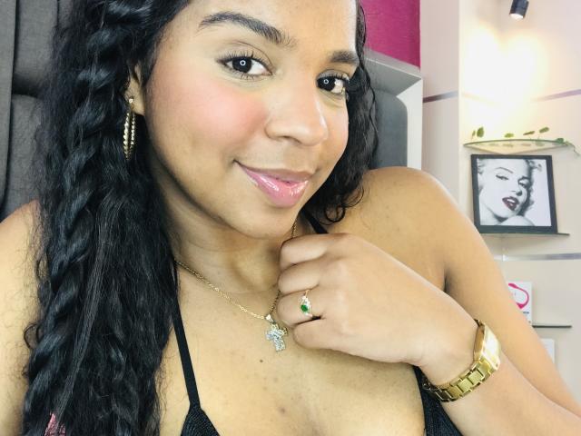 LisaMendez with curly black hair, pink glossy lips, gold hoop earrings, black halter top, gold watch, green ring, necklace, and a smile, showing her tits.